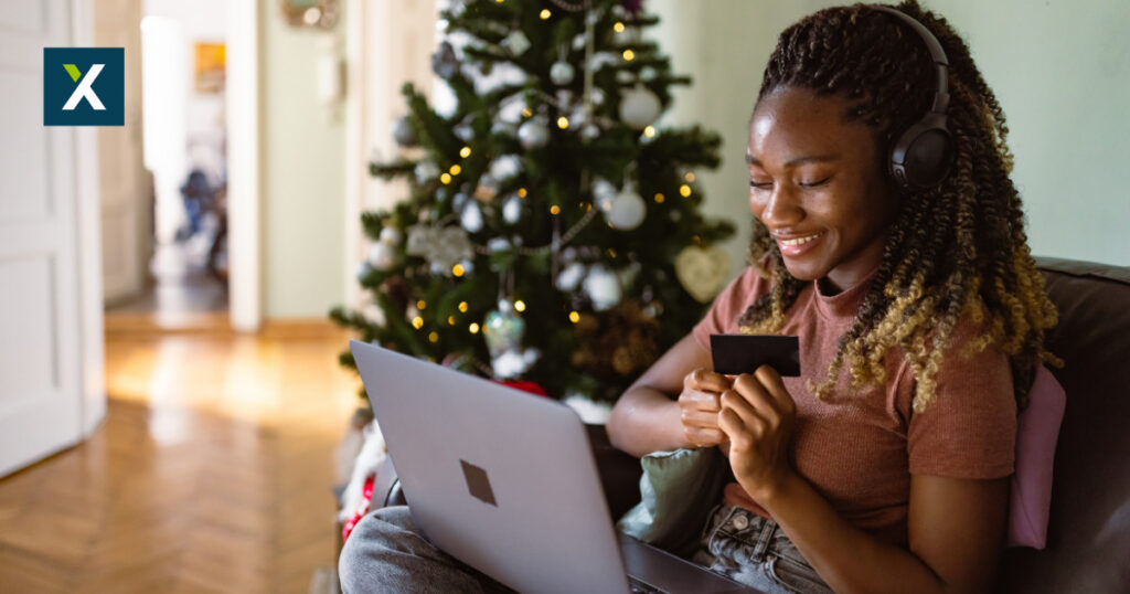 Online Shopperin sitzt mit dem Laptop auf dem Sofa. Im Hintergrund ist ein geschmückter Weihnachtsbaum.